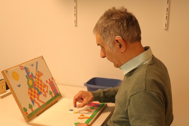 Bewoner met een hoortoestel achter het oor legt geconcentreerd een mozaïek aan een werktafel.